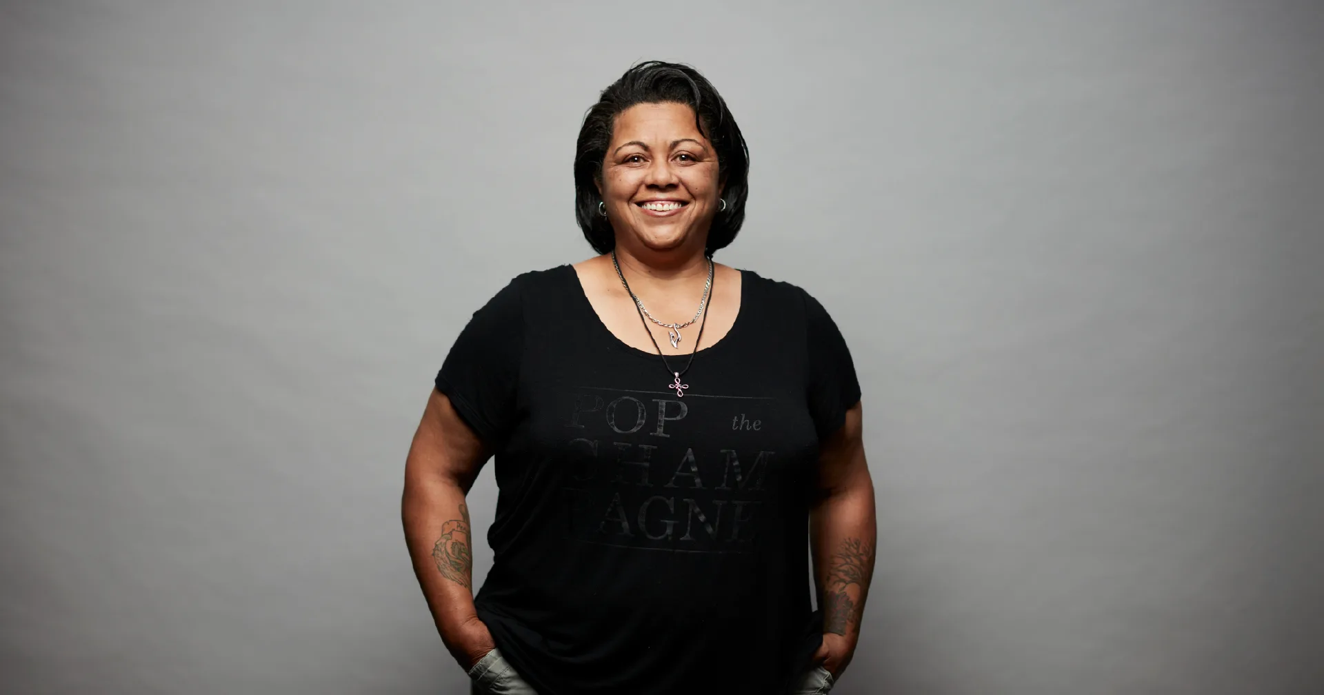 A person with short dark hair, smiling, stands against a plain gray background. They are wearing a black t-shirt with text and have their hands in their pockets. Visible tattoos are on both forearms.