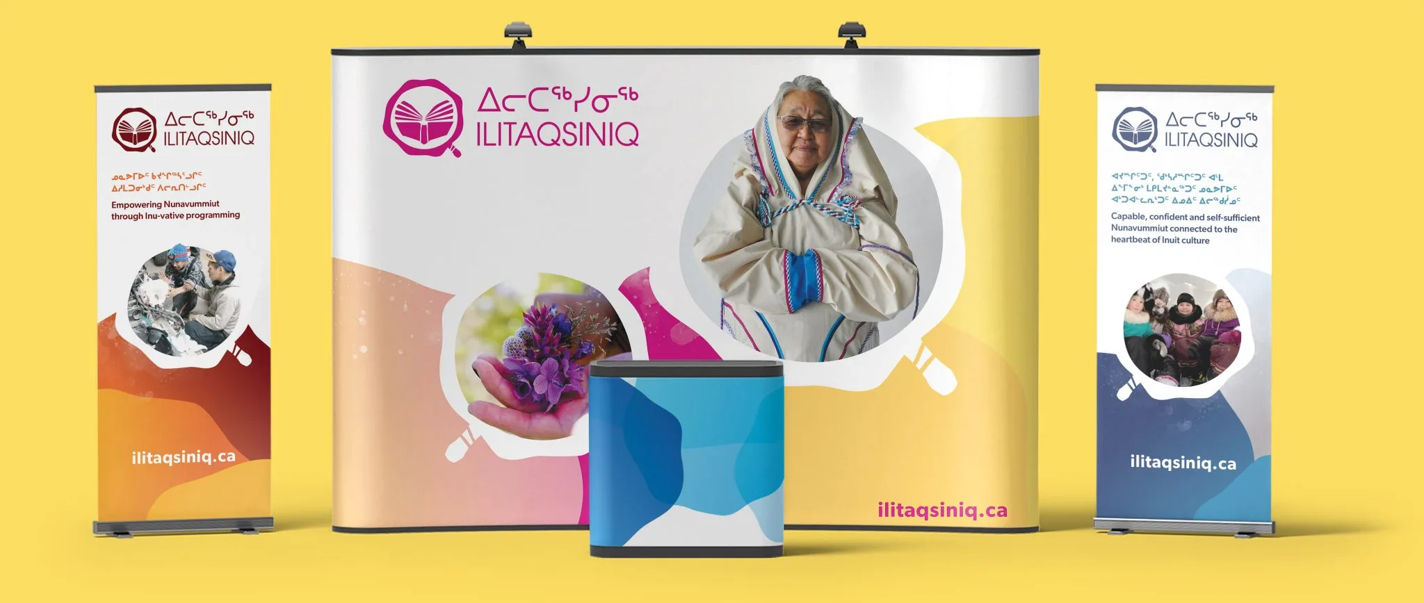 A trade show display with banners and a table promoting Ilitaqsiniq, featuring Inuktitut text, photos of Indigenous women and children, and images of traditional activities on a yellow background.