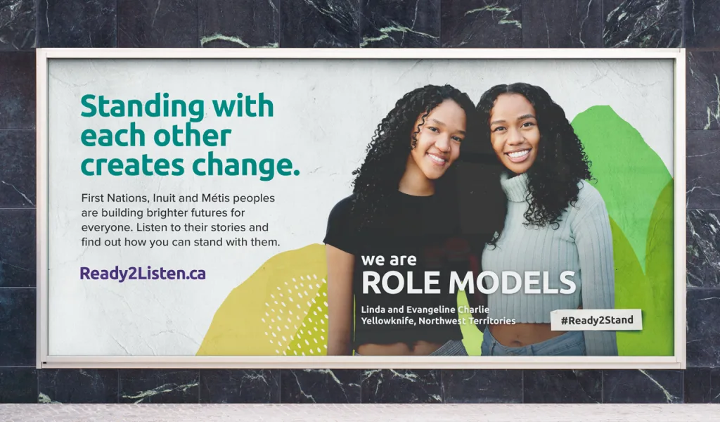 A billboard features two smiling women standing close together. Text reads: “Standing with each other creates change. We are role models. Linda and Evangeline Charlie, Yellowknife, Northwest Territories.” #Ready2Stand.