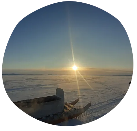 A wooden sled sits on a vast, snow-covered landscape under a clear sky, with the sun low on the horizon casting a warm glow across the icy scene.