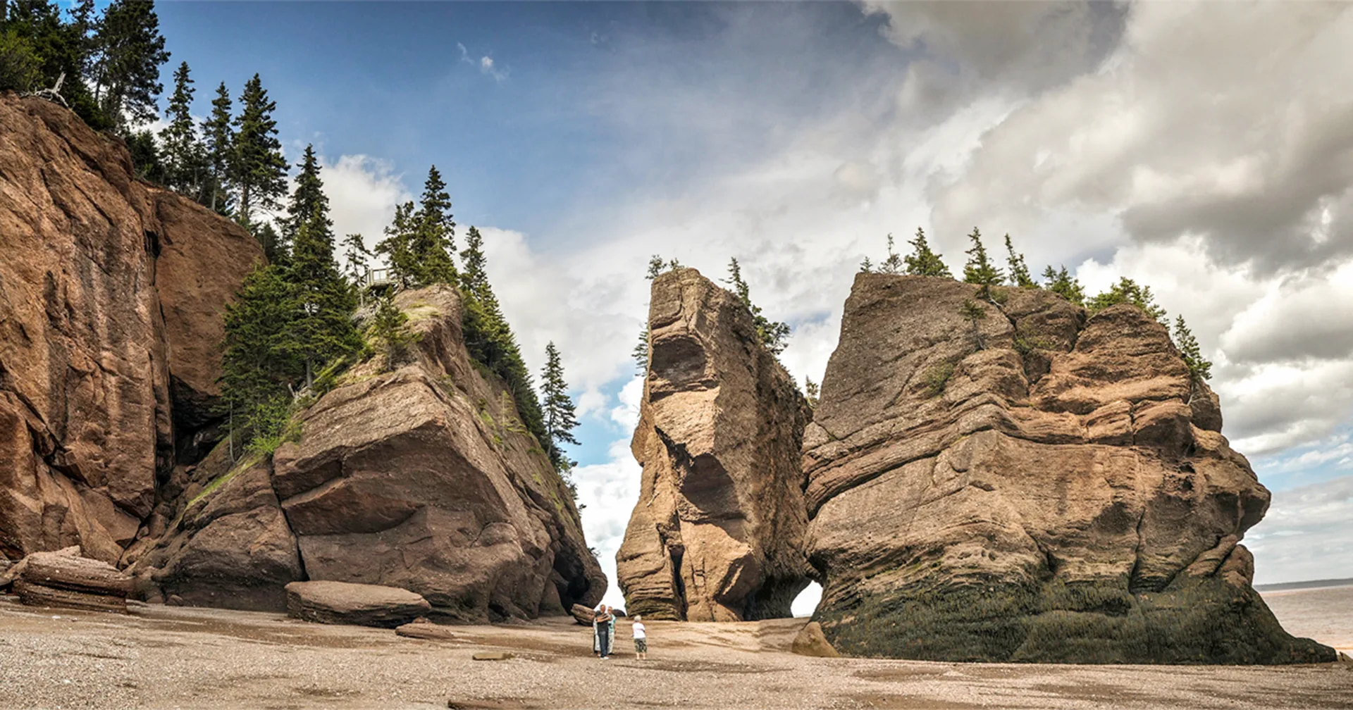 People stand near large, rocky sea stacks topped with trees at Hopewell Rocks on a sandy beach, with partly cloudy skies above and cliffs covered in greenery on the left.