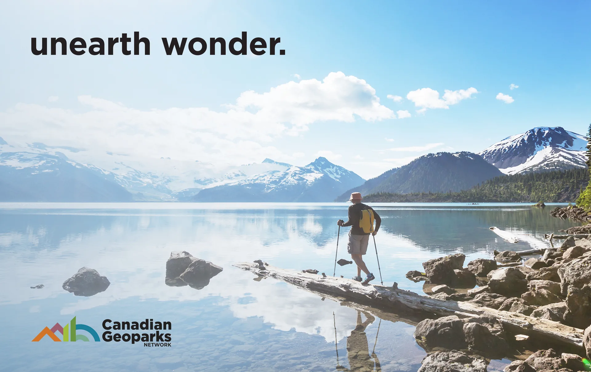 A hiker with trekking poles stands on rocks by a calm lake, surrounded by snow-capped mountains and pine trees under a blue sky. Text reads unearth wonder. with the Canadian Geoparks Network logo in the corner.