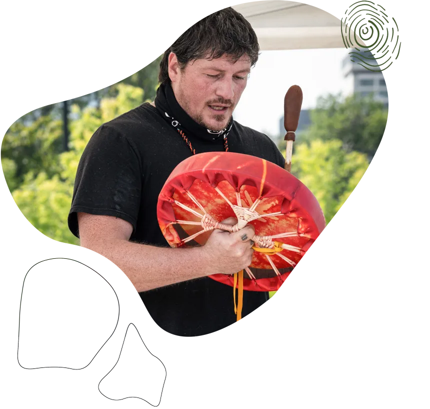 A man plays a colorful hand drum outdoors, holding a mallet and looking down with focus. Green trees and a building are visible in the blurred background. Abstract shapes decorate the image edges.