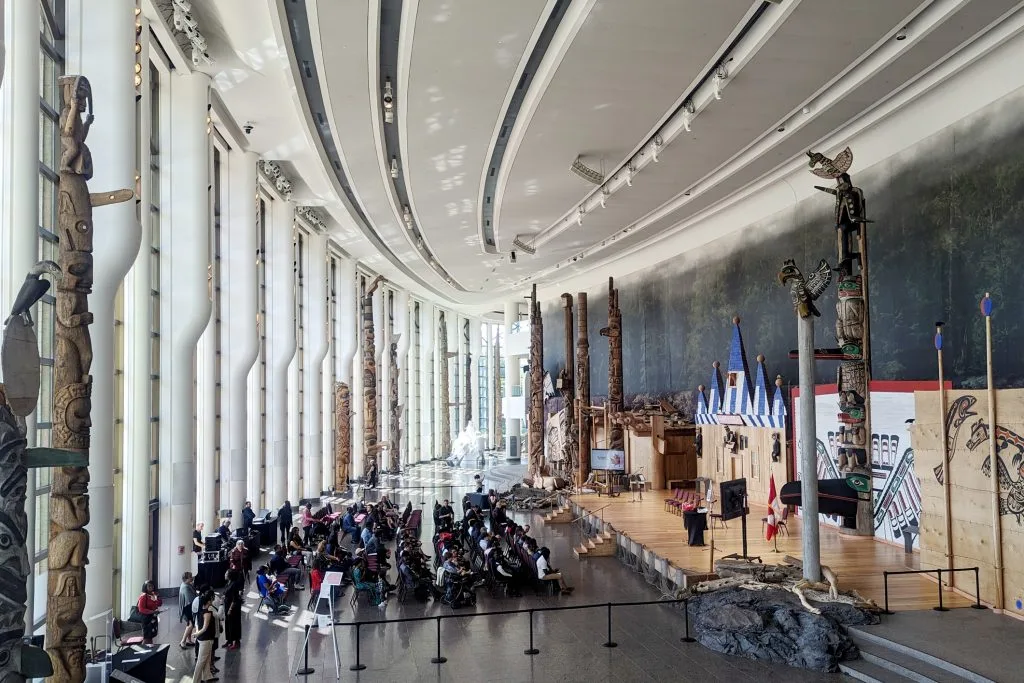 A spacious, modern hall with tall windows, Indigenous totem poles, and a wooden stage; people are seated and gathered along one side, bathed in natural light.