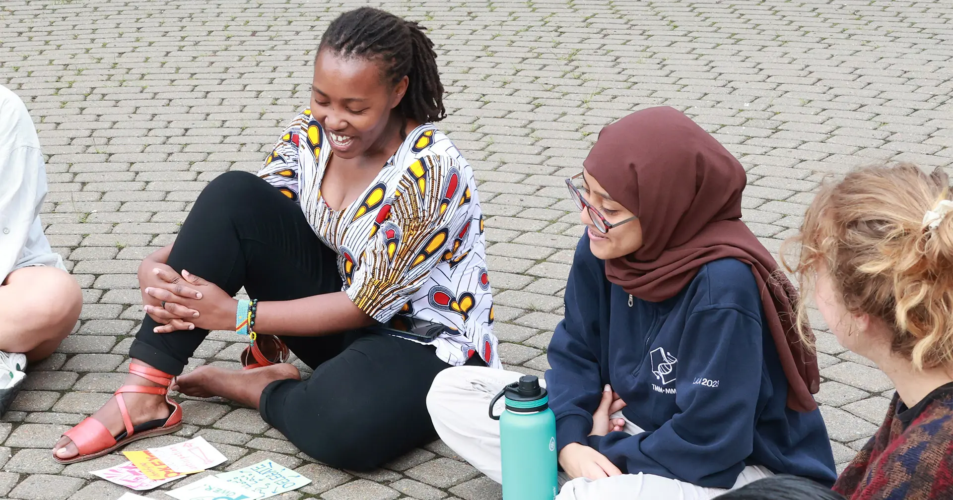 Three people sit on a paved surface, smiling and talking. One wears a colorful shirt and sandals, another wears a brown hijab and glasses, and the third has light brown hair. Papers and a teal water bottle are on the ground.