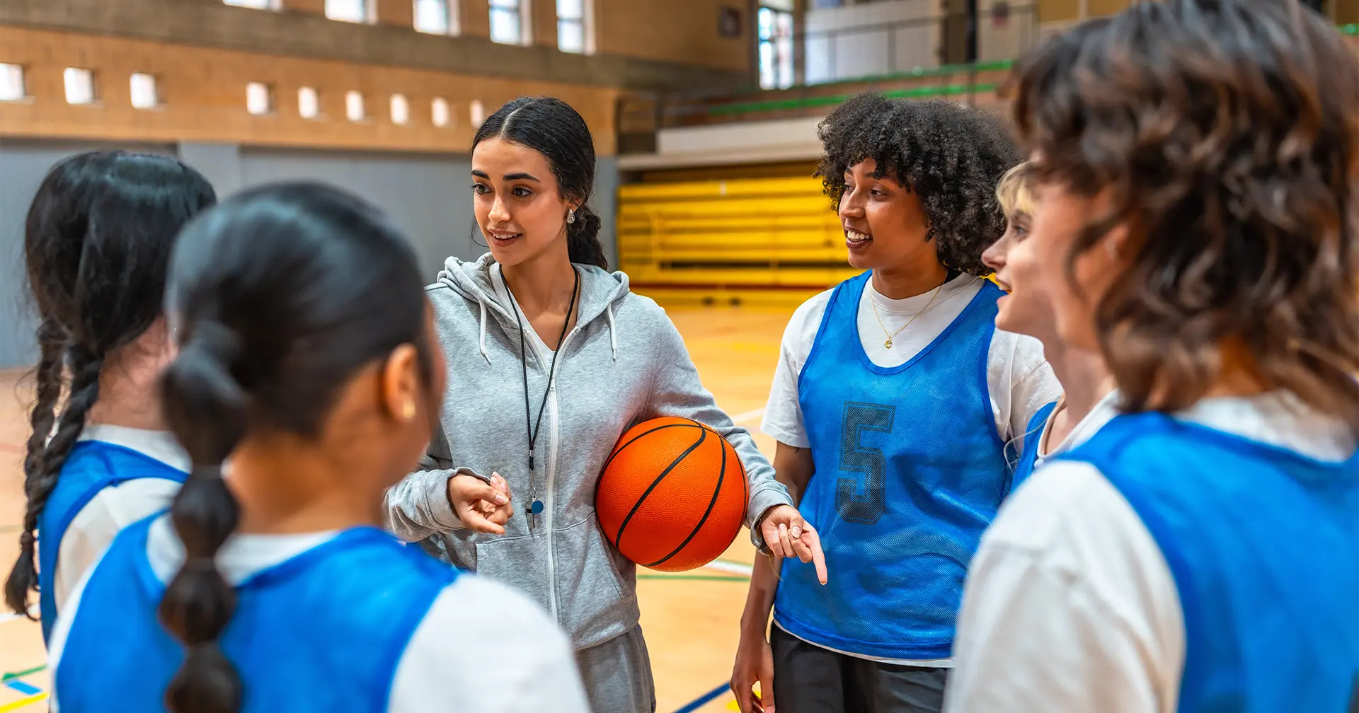 A basketball coach holding a ball gives instructions to four young players wearing blue jerseys inside a gymnasium. The team stands in a huddle, listening attentively.