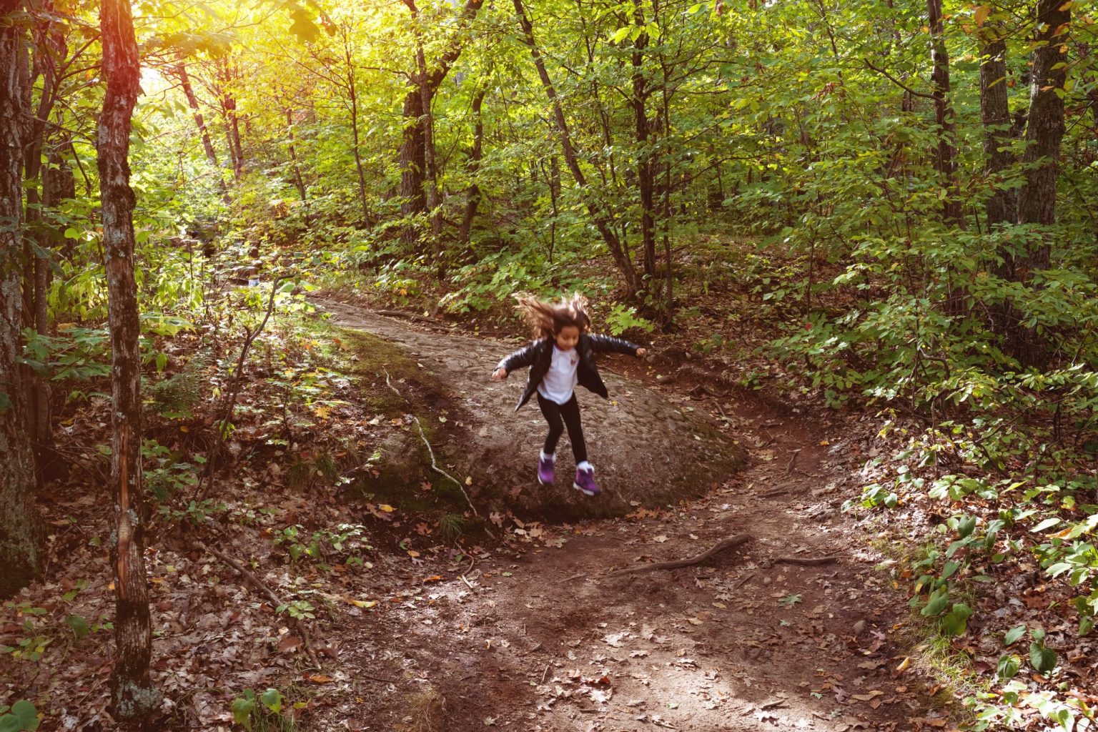 A young girl in a jacket jumps off a small incline on a forest trail, surrounded by lush green trees and sunlight filtering through the leaves.