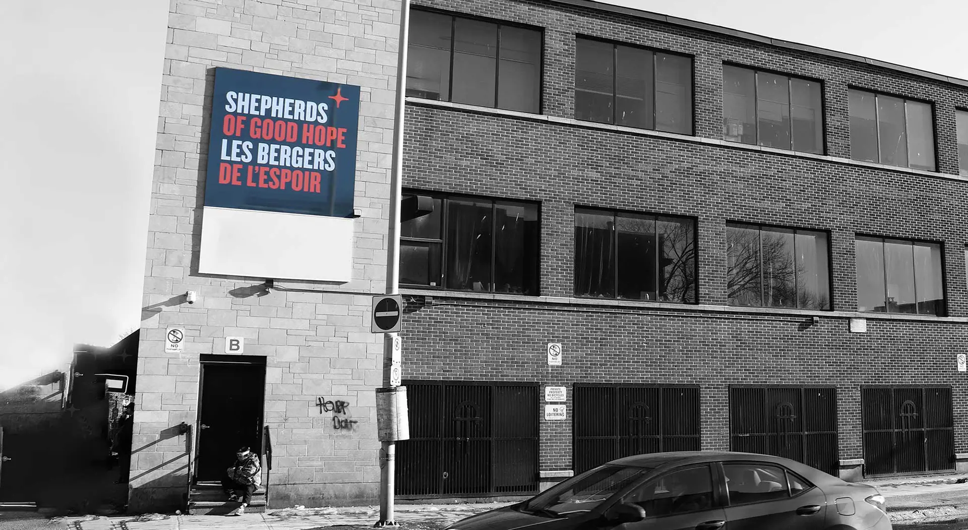 A brick building with a bilingual sign reading Shepherds of Good Hope / Les Bergers de L’Espoir in red and white on blue, above a person sitting by the entrance on a snowy sidewalk next to a parked black car.