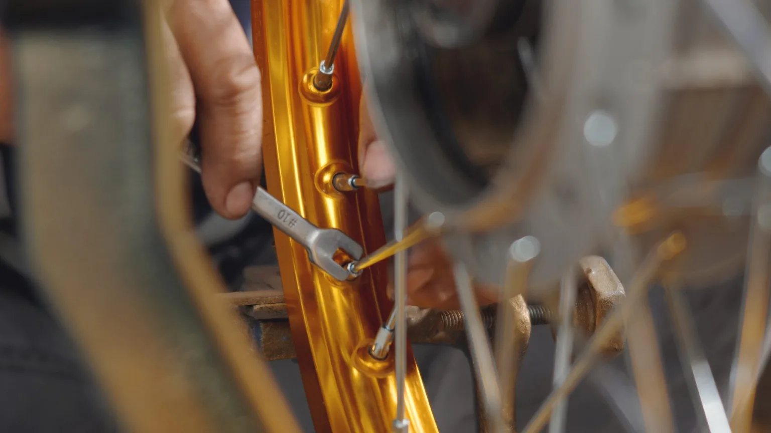 A person uses a spoke wrench to tighten the spokes on an orange bicycle or motorcycle wheel rim, performing maintenance or repairs. The image is a close-up, showing hands, spokes, and part of the wheel.