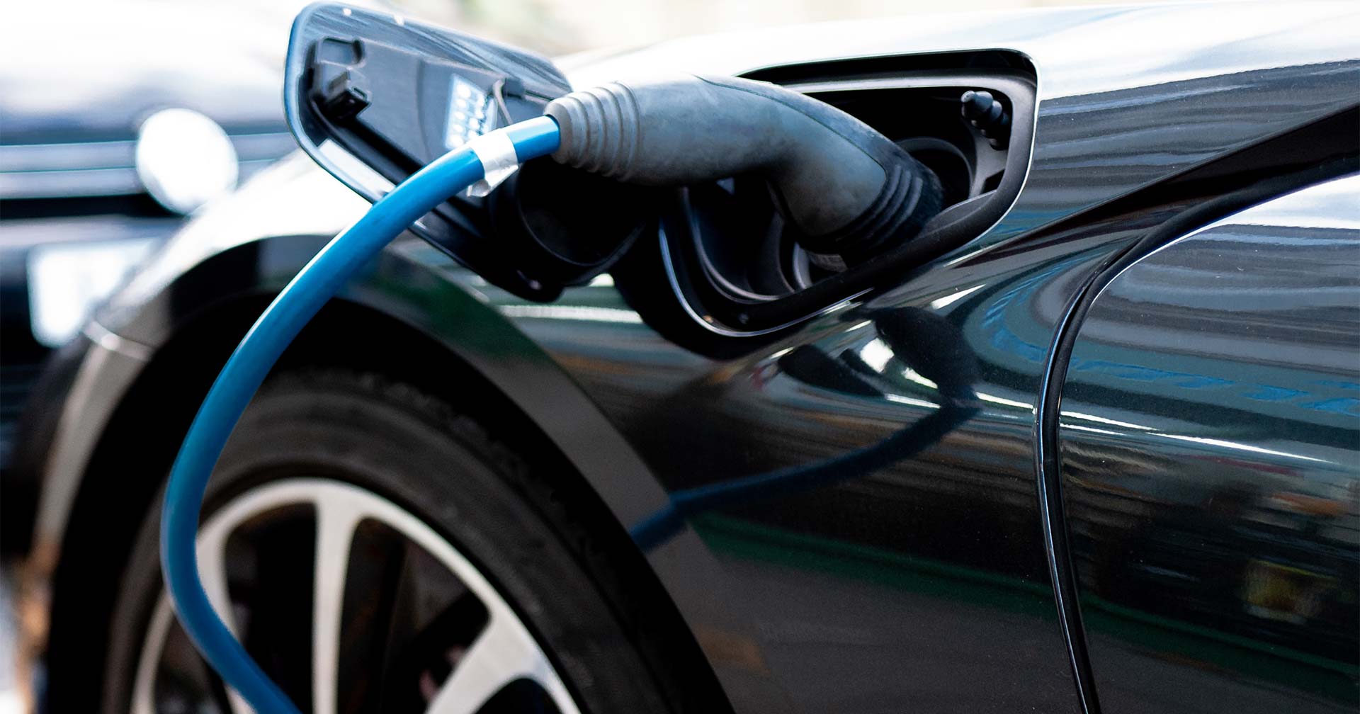 Close-up of an electric vehicle being charged, with a blue charging cable plugged into the cars charging port near the front wheel. The shiny black car reflects its surroundings.