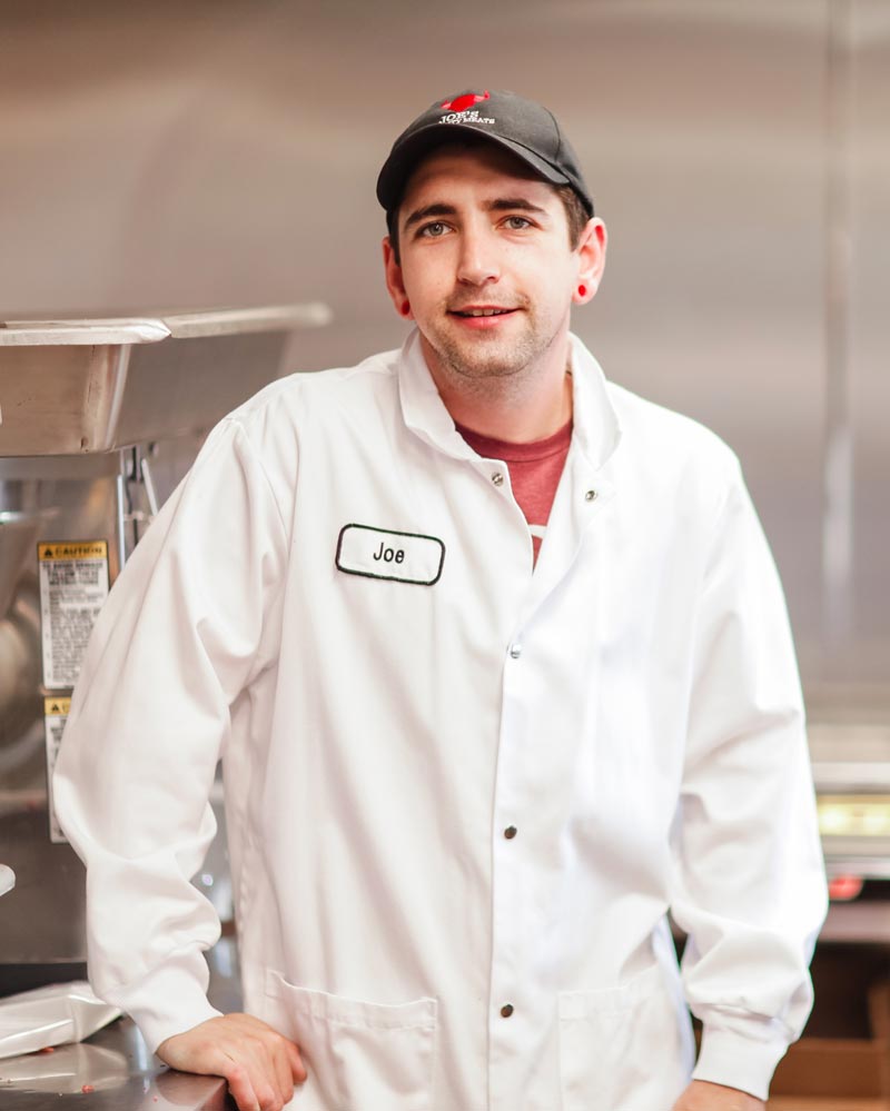A man in a white lab coat and black cap, with a name tag that says Joe, stands in a commercial kitchen, smiling slightly with one hand resting on the counter.