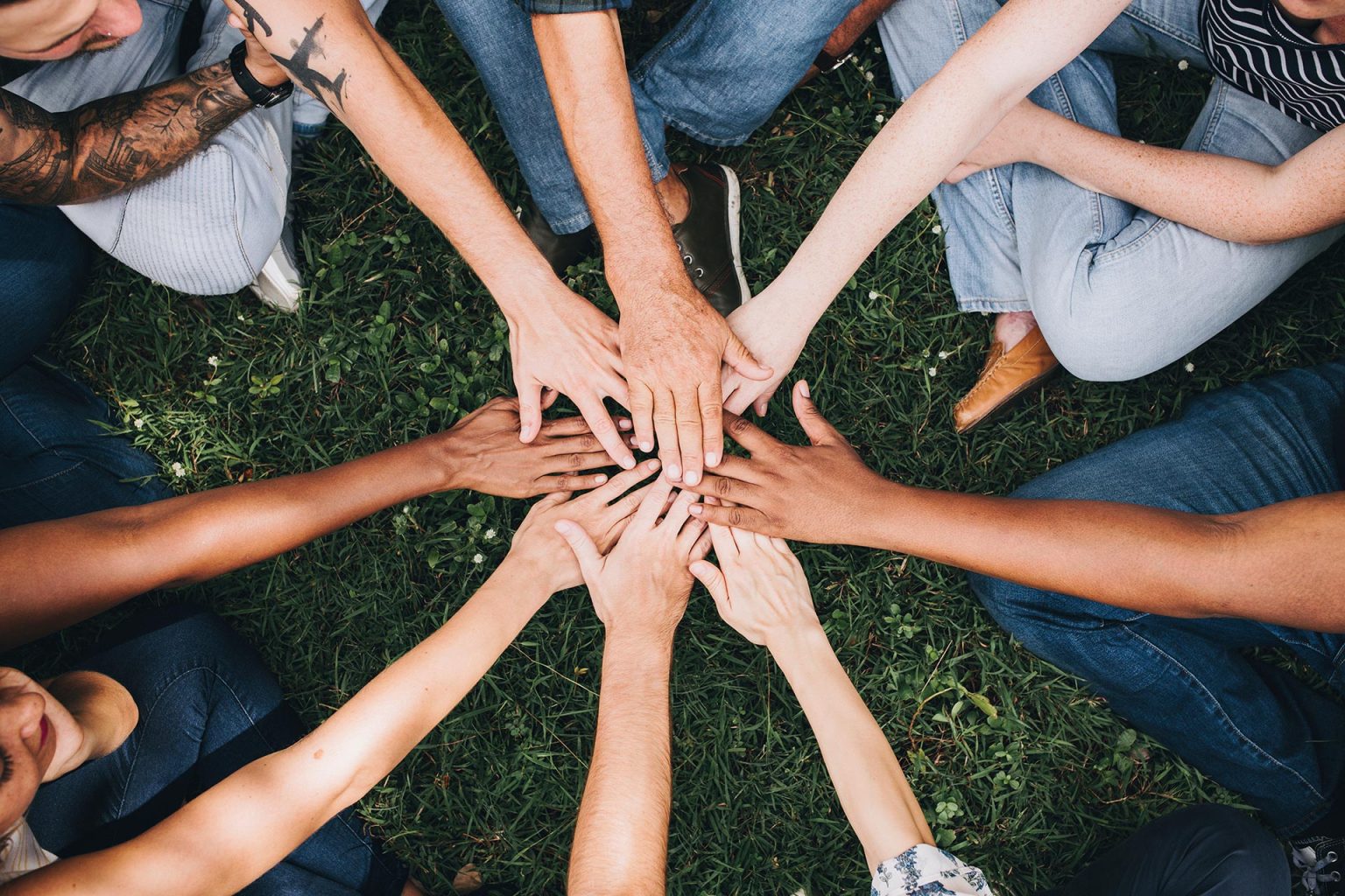 A group of diverse individuals sit in a circle and reach to the middle to hold their hands together.