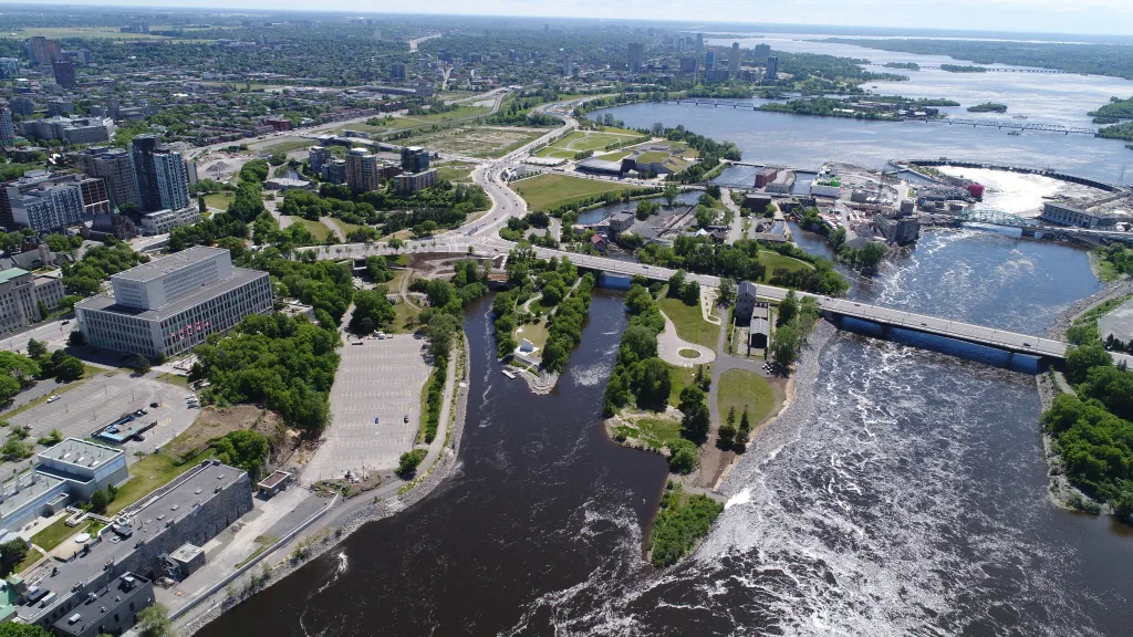 Aerial view of a city with a river running through it, green parks, several bridges, and buildings along the water’s edge under a partly cloudy sky.