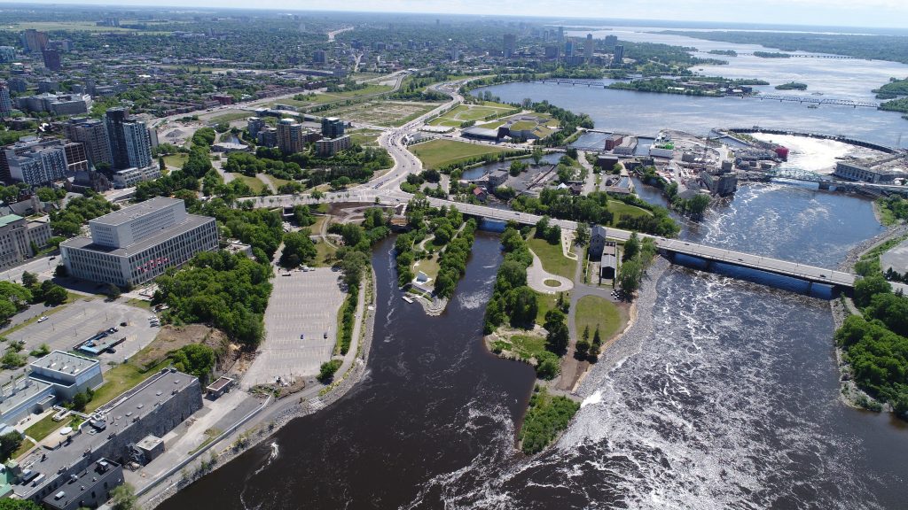 Aerial view of a city with a river running through it, green parks, several bridges, and buildings along the water’s edge under a partly cloudy sky.