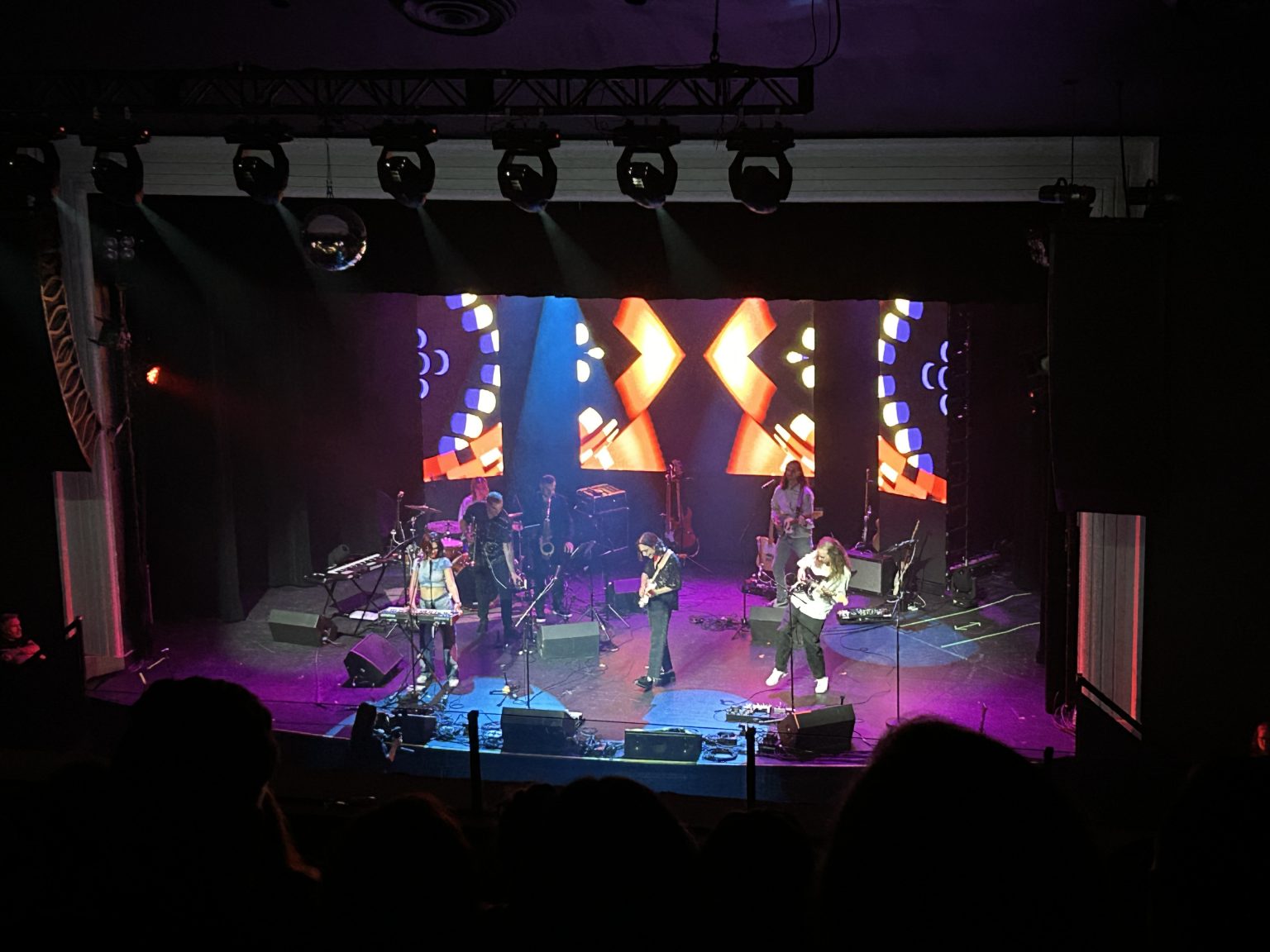 A band performs on stage with colorful lights and abstract visuals on a large screen behind them, while the audience watches from darkened seats in a theater.