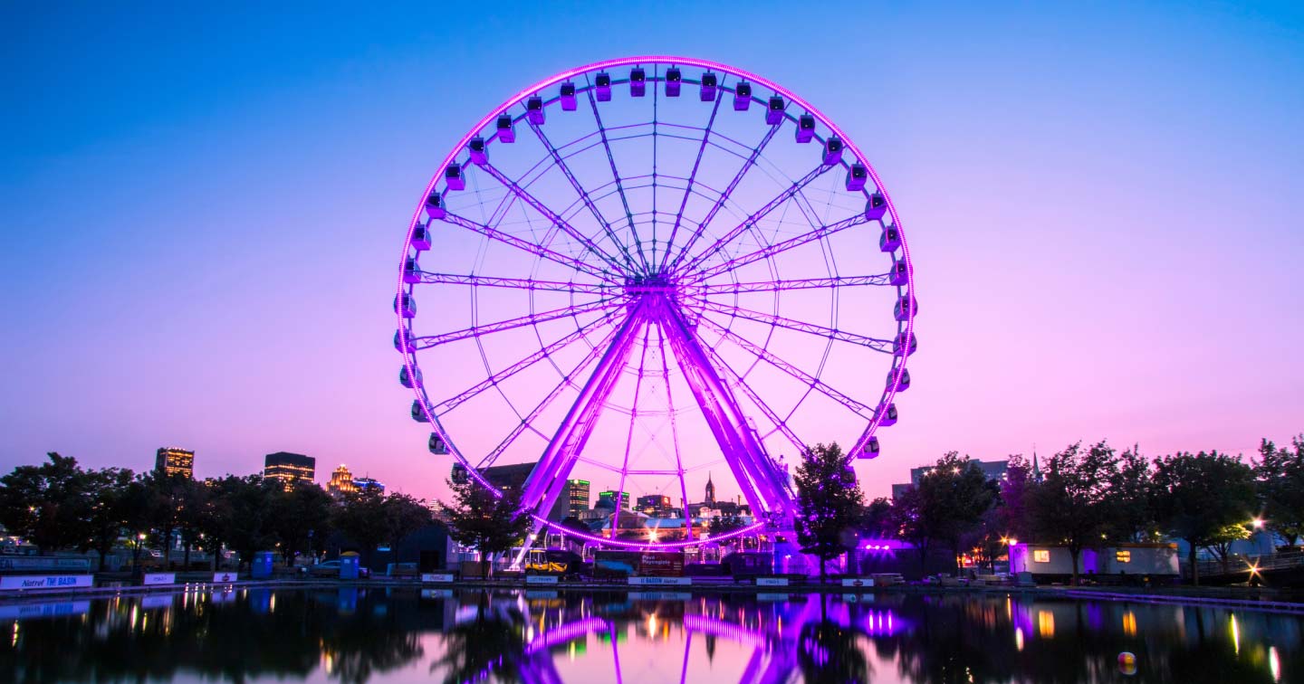 A large Ferris wheel glowing with purple lights stands beside a reflective pond at dusk, with city buildings and trees silhouetted against a vibrant pink and blue sky.