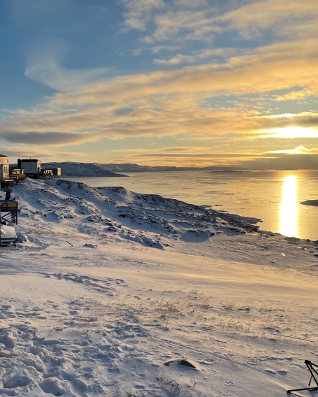 Snow-covered coastline at sunset, with houses on the left, rocky shoreline, and calm water reflecting the golden sky and sun. Thin clouds streak across the colorful sky, creating a peaceful winter scene.