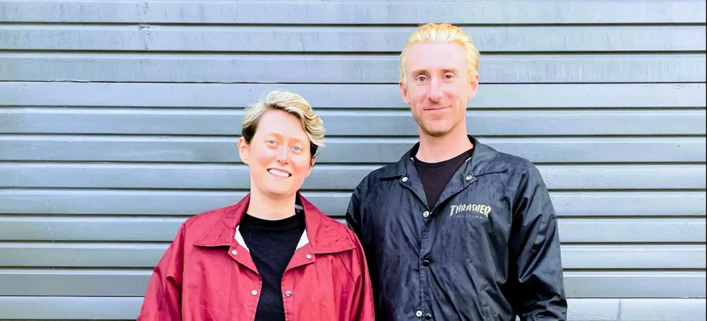 Two people with short blonde hair stand side by side smiling in front of a gray metal garage door. One wears a red jacket, and the other wears a black jacket over a black shirt.