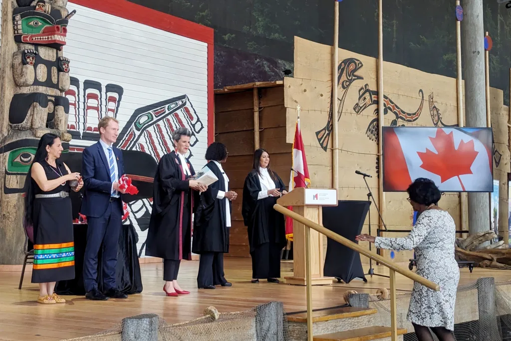 People on stage at a citizenship ceremony, with officials in robes and a woman approaching to be sworn in. A large Canadian flag is displayed on a screen, and Indigenous art decorates the background.