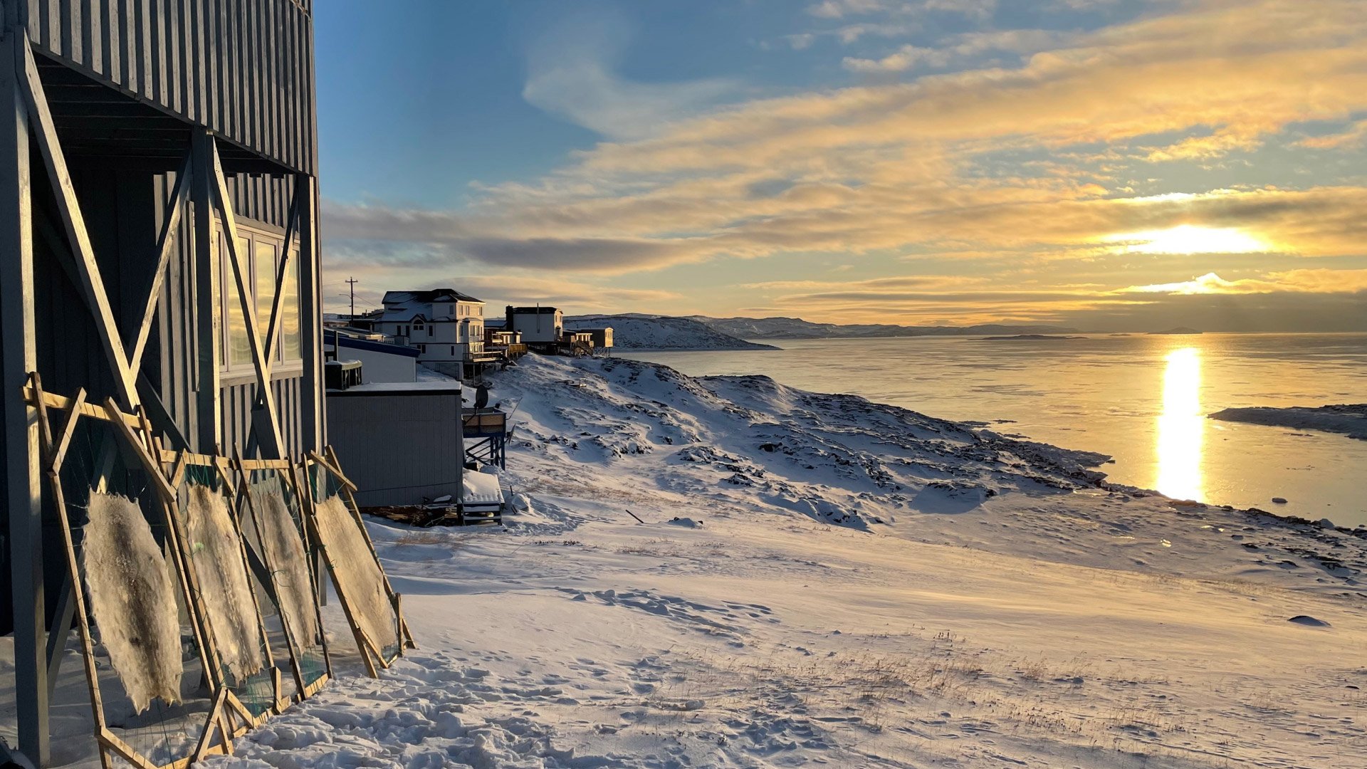 A snowy coastal village at sunset, with houses on stilts along the shoreline and animal skins stretched on wooden frames outside. The sun reflects on calm, icy water under a partly cloudy sky.
