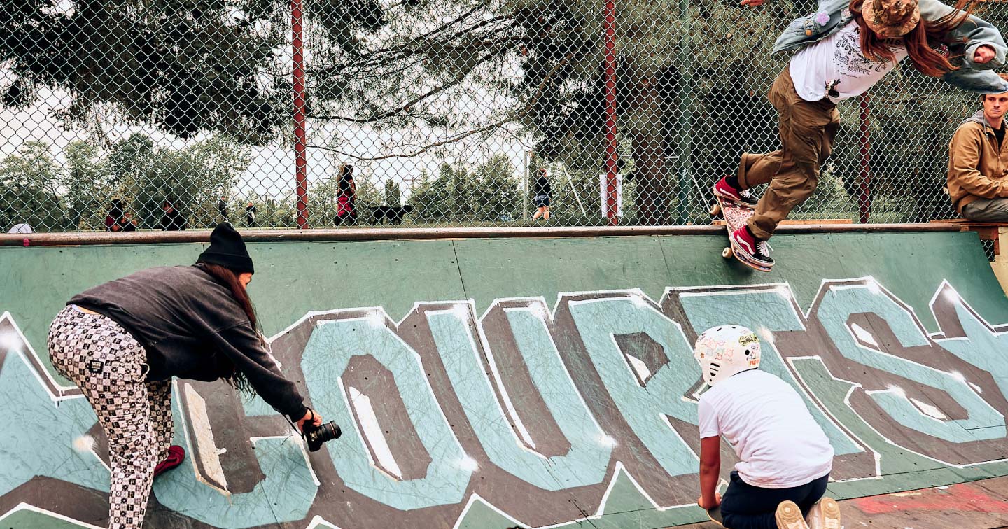 Three people at a skatepark: one person skateboards on a graffiti-covered ramp, another films them with a camera, and a third, wearing a helmet, kneels nearby. Trees and a chain-link fence are in the background.
