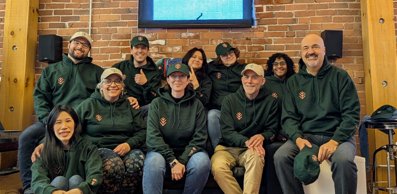 A group of eleven people sit and kneel together indoors, smiling and wearing matching dark green hoodies and hats. They pose casually against a brick wall with a TV screen above them.