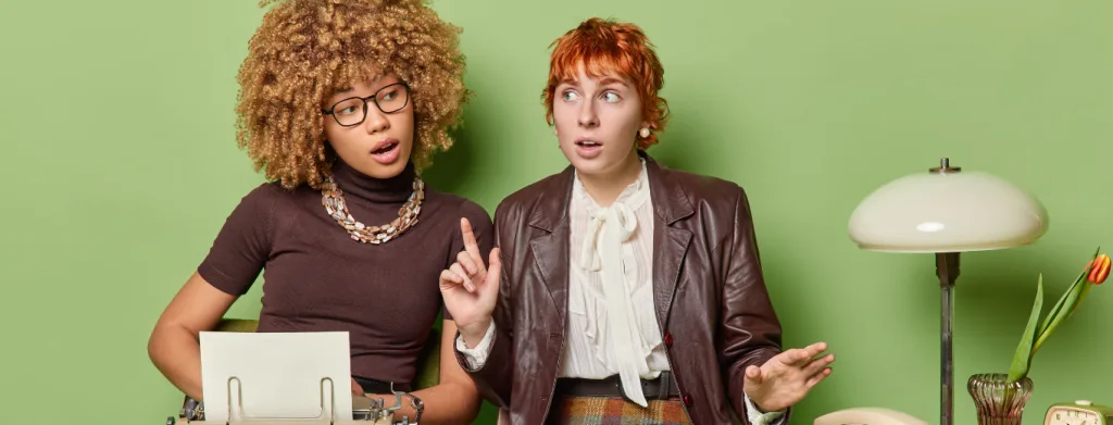 Two women sit at a desk with a typewriter and lamp against a green background. One woman with curly hair and glasses looks thoughtful, while the other with short red hair gestures as if explaining something.
