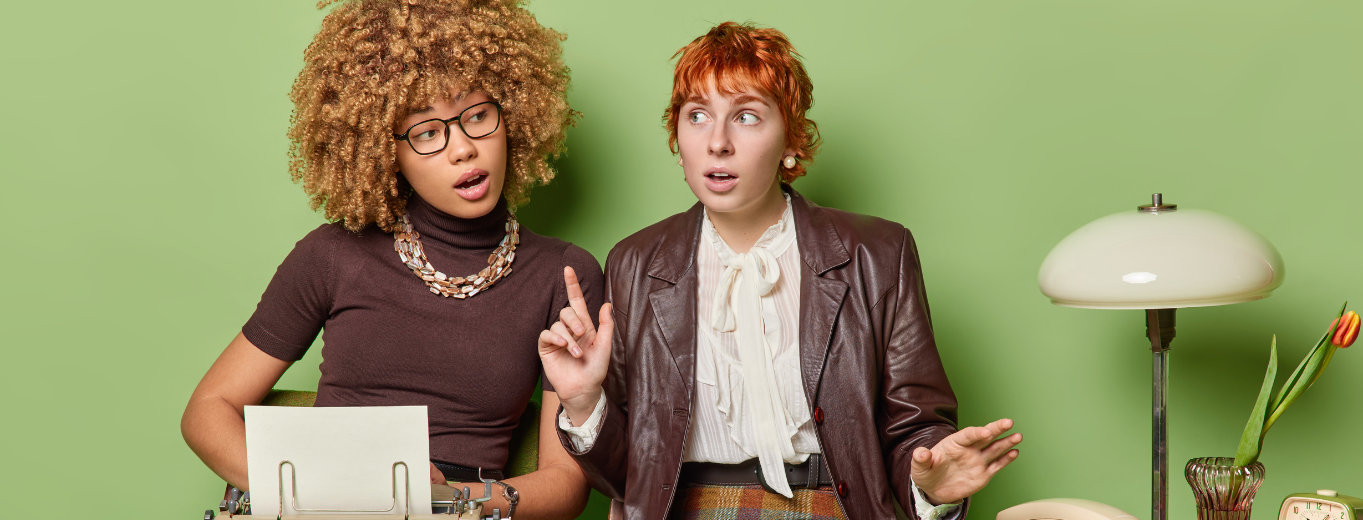 Two women sit at a desk with a typewriter and lamp against a green background. One woman with curly hair and glasses looks thoughtful, while the other with short red hair gestures as if explaining something.