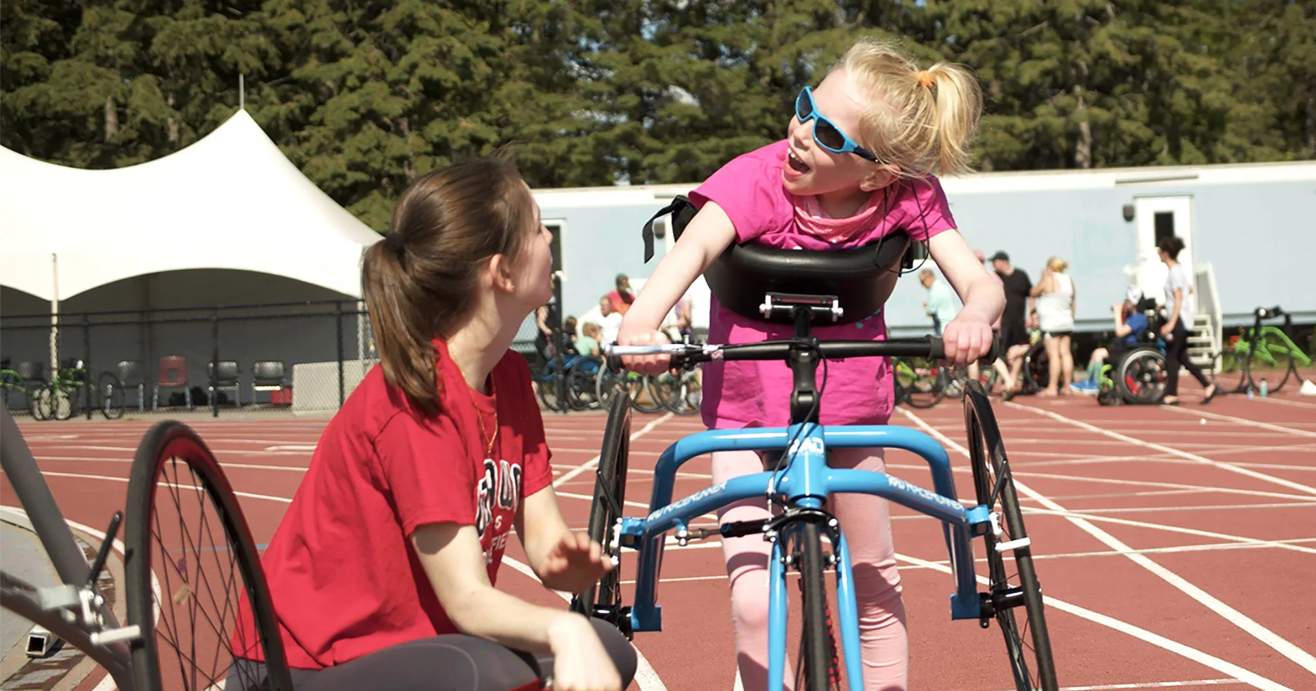 A young girl using a blue adaptive tricycle smiles and talks to a woman in a red shirt on a running track, with trees and people in the background.