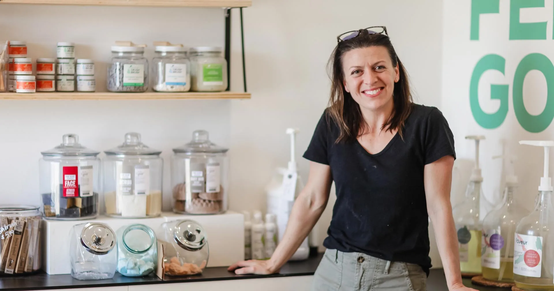 A woman wearing a black t-shirt and khaki pants smiles while standing in a store with shelves of jars containing various products and bottles. The wall behind her has green letters partially spelling “FEEL GOOD.”.