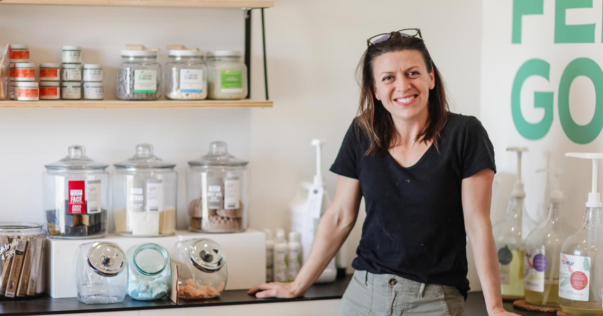 A woman wearing a black t-shirt and khaki pants smiles while standing in a store with shelves of jars containing various products and bottles. The wall behind her has green letters partially spelling “FEEL GOOD.”.
