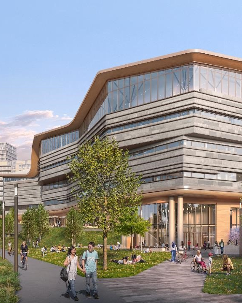 A modern, multi-story building with large glass windows and curved lines sits beside a green lawn where people are walking, cycling, and relaxing under a blue sky.