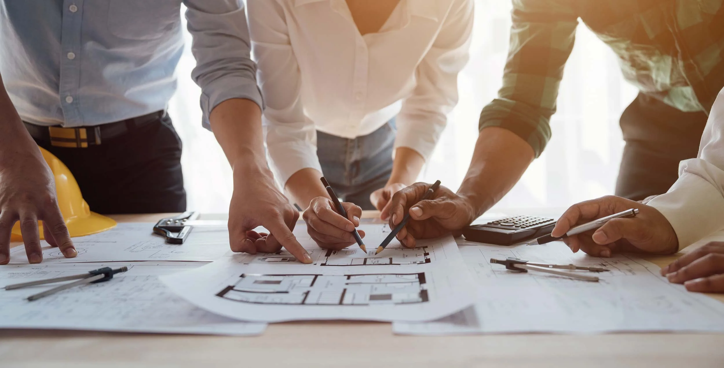 Several people standing around a table collaborate on architectural blueprints, using pens and compasses, with a yellow hard hat and calculator visible nearby.