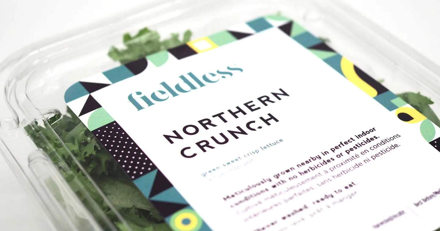 A plastic container labeled Fieldless Northern Crunch holds green sweet crisp lettuce. The label highlights that the lettuce is grown without herbicides or pesticides and features a colorful geometric border.