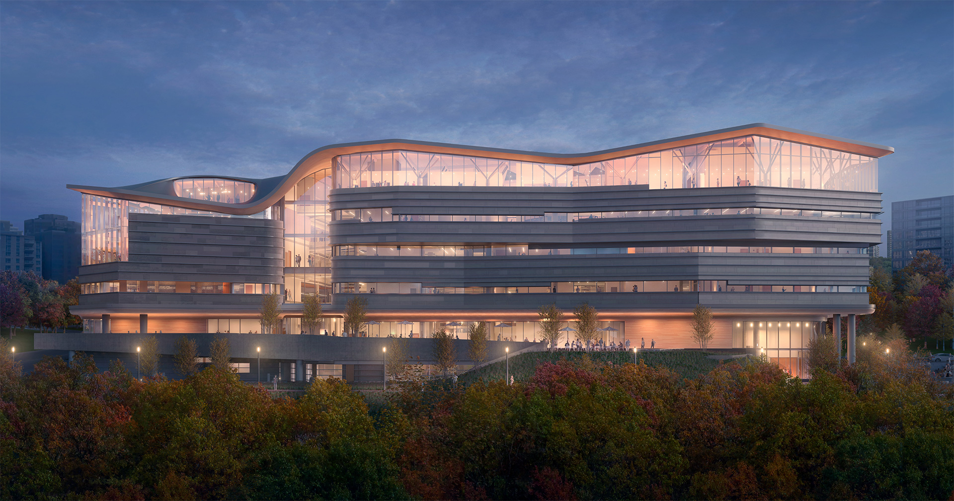 A modern, multi-story building with large glass windows is illuminated at dusk, surrounded by trees with autumn foliage, and set against a cloudy evening sky.