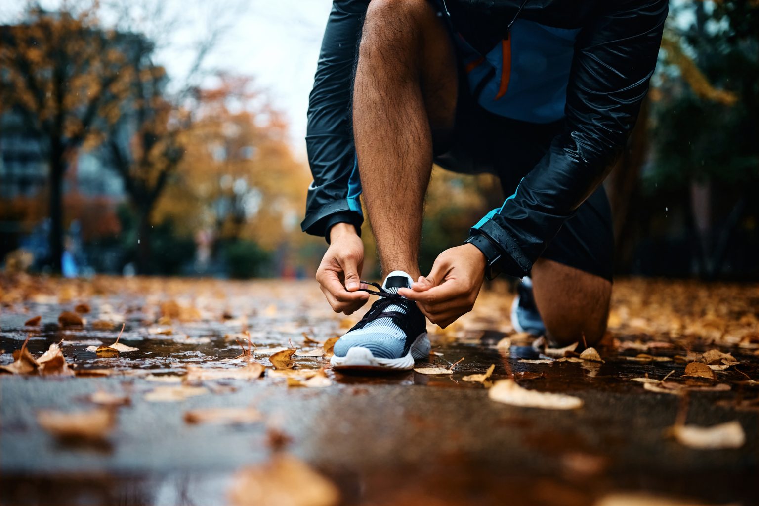 A person in athletic wear kneels to tie their running shoe on a wet, leaf-covered path in a park during autumn. The scene is outdoors and the ground is damp with fallen leaves.