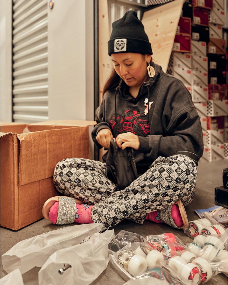 A person in a beanie, hoodie, and patterned pants sits cross-legged on the floor, packing items into bags. Open boxes and various small objects, like ping pong balls, are scattered around them.