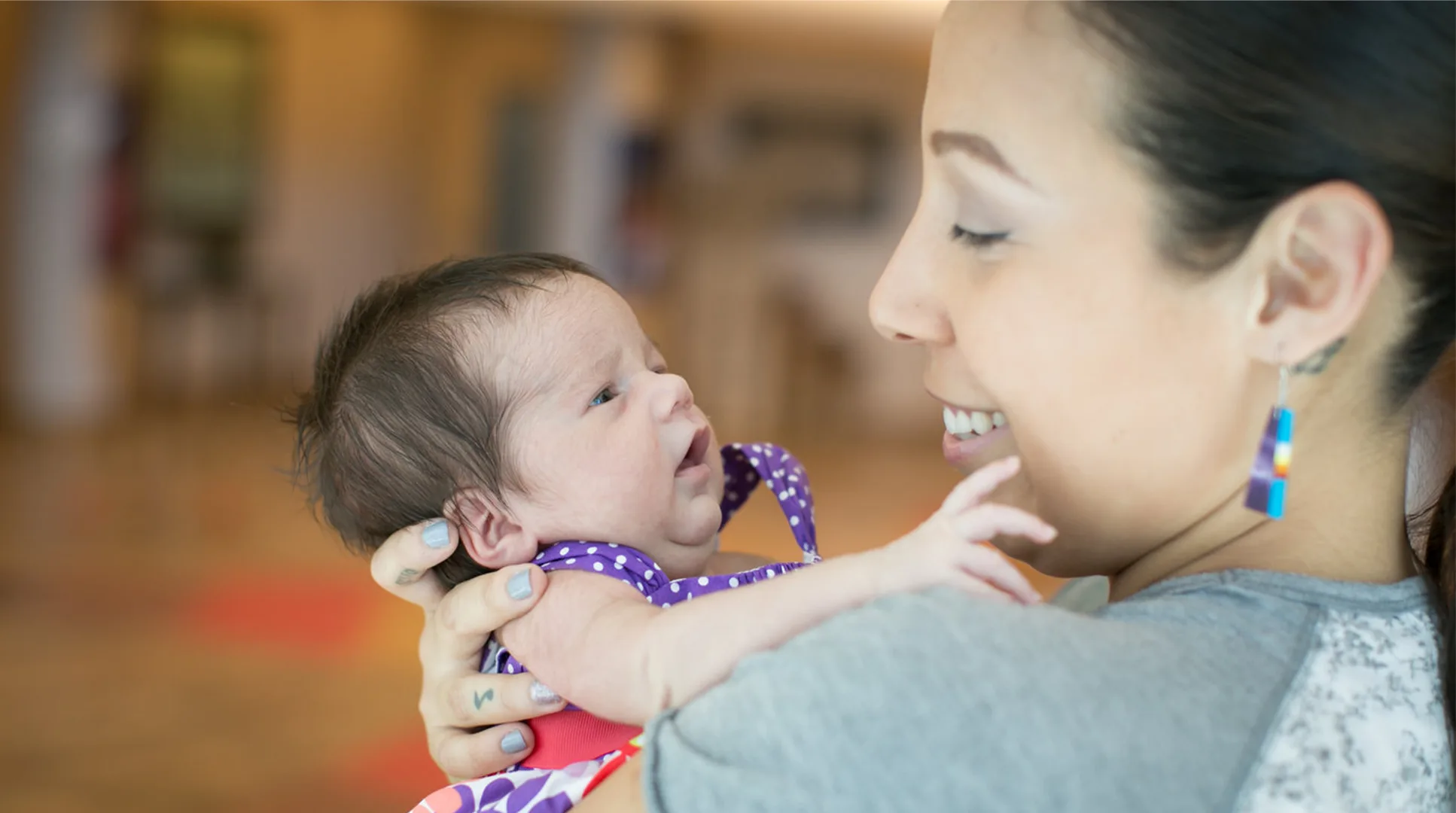 A woman smiling and holding a newborn baby dressed in a purple outfit. The baby looks up at the woman. The background is softly blurred.